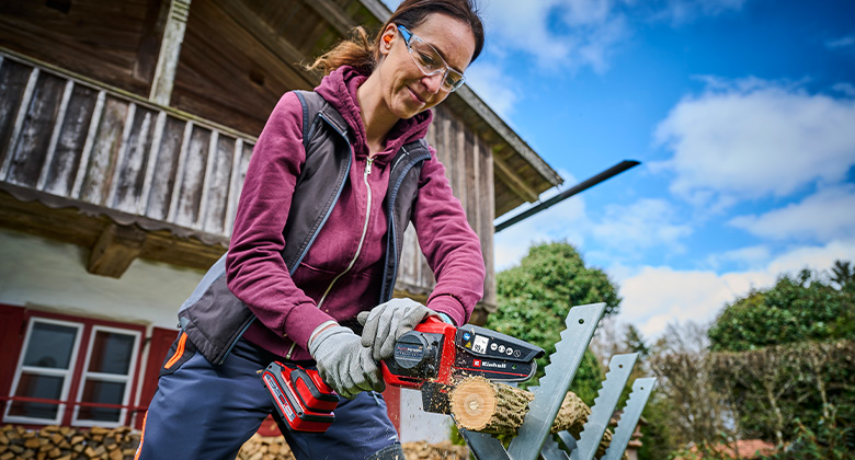 A woman wearing safety goggles saws a small log outdoors with an Einhell chainsaw.