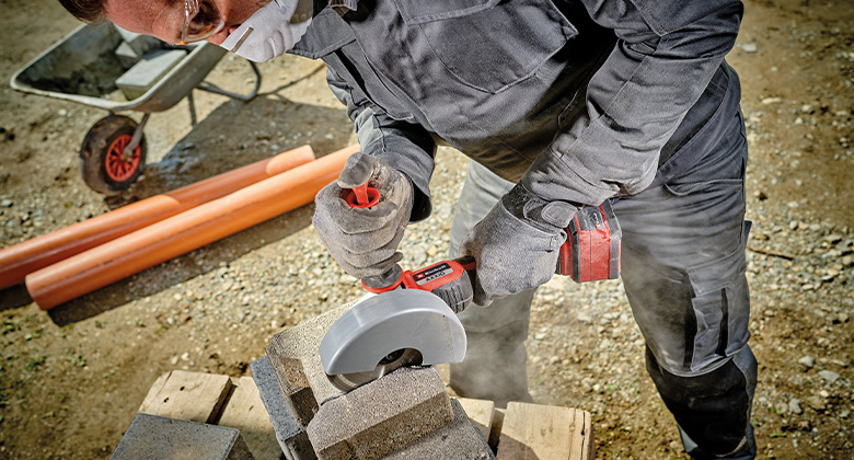 A person cuts concrete blocks with an Einhell cordless cut-off grinder, with dust rising.