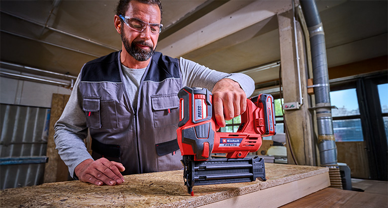 A man is using an Einhell cordless nailer FIXETTO to work on a wooden piece, wearing safety glasses and workwear.