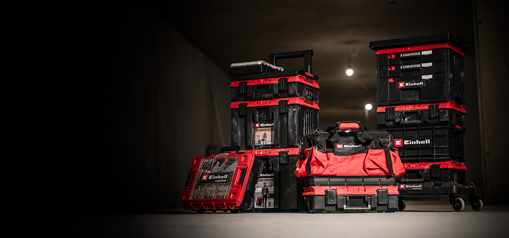 Several black and red Einhell E-Case toolboxes and bags are stacked and arranged in a dark room.