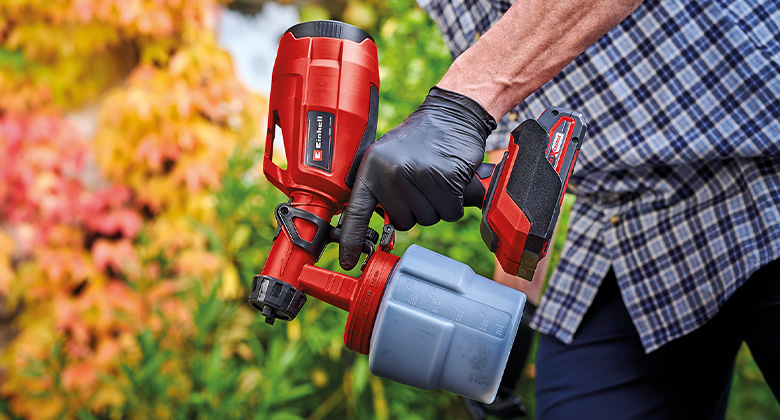 A person in a checkered shirt holds a paint sprayer while wearing a black glove.