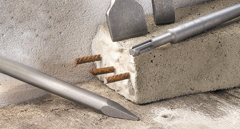Various chisel attachments are placed next to a concrete block with exposed metal rods.