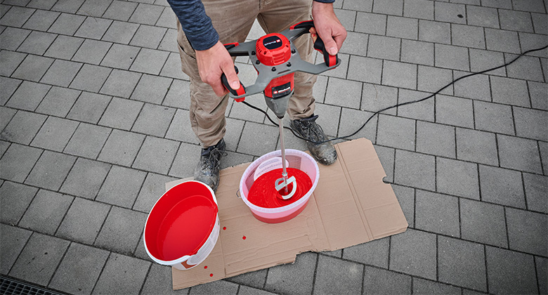 A person uses a mixer to stir red paint in a bucket placed on cardboard.