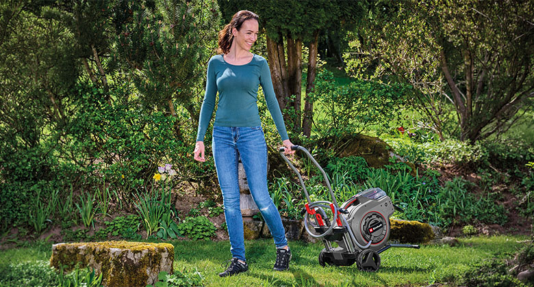 Woman pushes a compact lawn mower with a black and red housing across a green lawn in a garden.