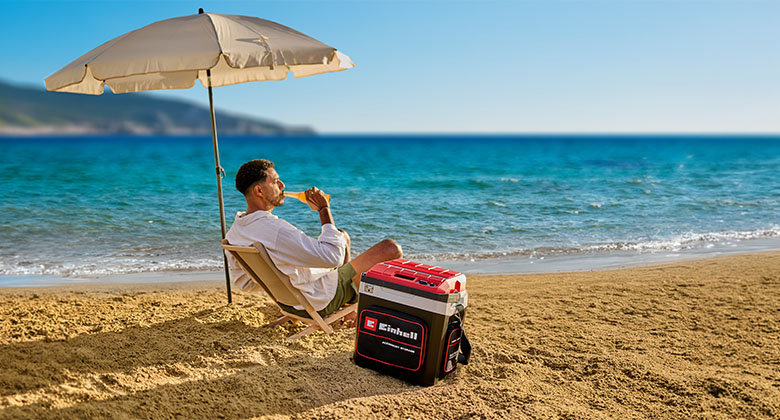 Man sitting on the beach under a parasol next to an Einhell cooler with red lid by the sea.