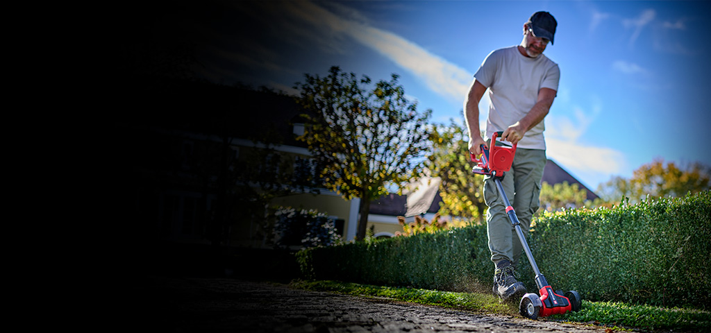 A man cleans paving joints along a hedge in the garden using the Einhell cordless patio cleaner.