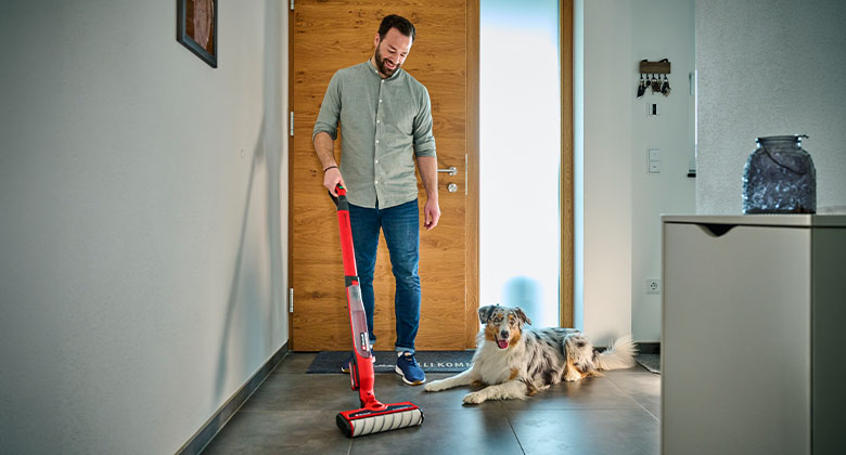 Man cleaning the hallway with a red cordless hard floor cleaner, a dog lying on the floor beside him.