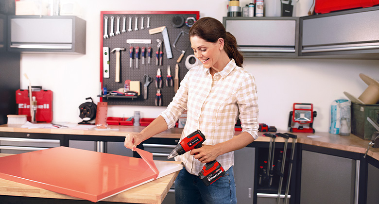 Woman uses a red heat gun to remove a pink film from a wooden board in a workshop.