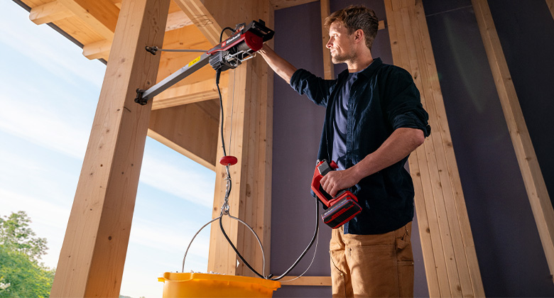 Man installing a cordless hoist on a wooden beam structure and lifting a yellow bucket.