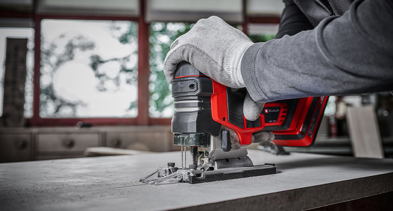 Gloved hand guiding a red cordless jigsaw through a wooden board.