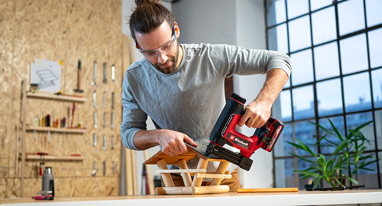 Man wearing safety glasses fastening wooden pieces with a red cordless nailer on a workbench.